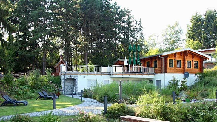 Foto zeigt Blockhaus mit Terrasse im Ferienpark Hesselhof. Dahinter (rechts im Bild) Gebäude mit Ferienwohnungen. Vor der Terrasse Liegewiese mit Liegen.